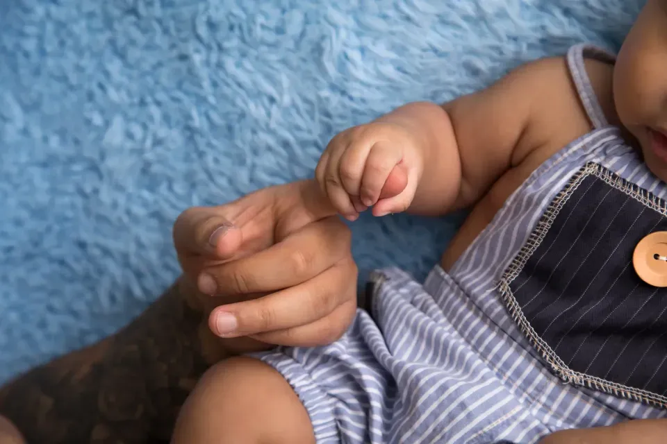 Newborn baby grasping an adult's finger against a blue textured background, wearing light-colored clothing.
