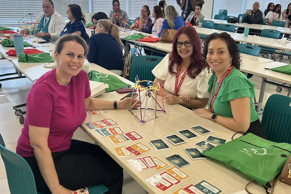 Three smiling women seated at a table with a colorful geometric building structure and educational cards, during a team-building workshop.